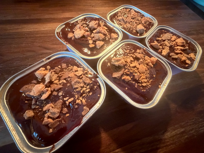 Five chocolate-topped dessert tins covered with golden Butterfinger pieces on a wooden board